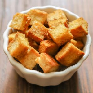 Close-up of golden-brown croutons in a white ceramic bowl on a rustic wooden table.