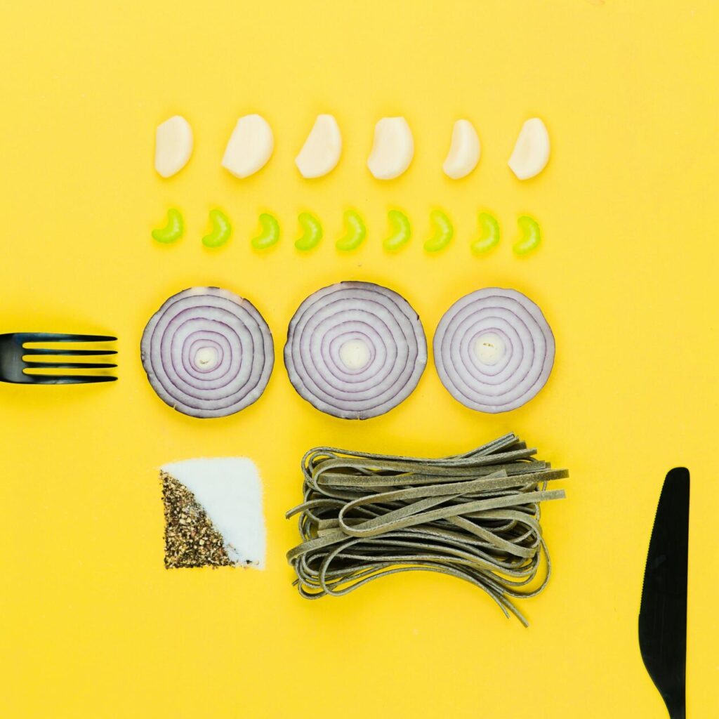 A colorful flatlay of pasta, onion, garlic, herbs, and cutlery on a yellow background.