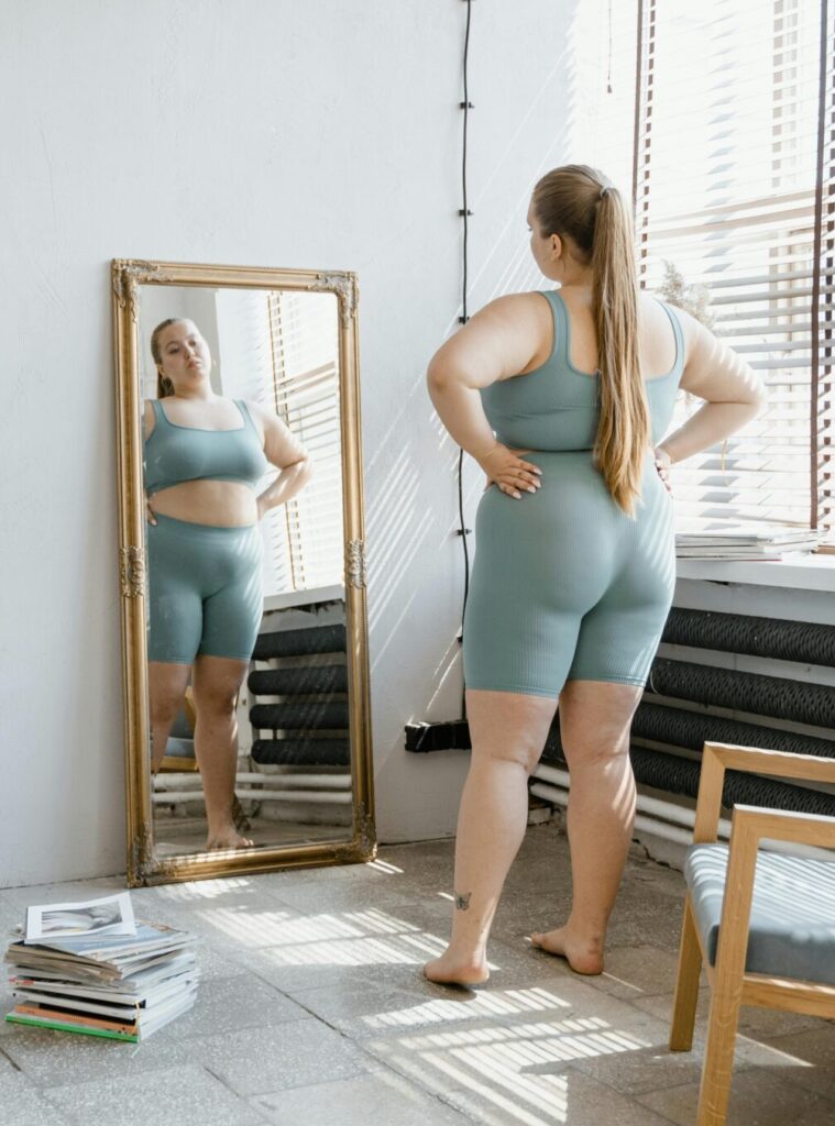 Plus size woman in sportswear stands barefoot, looking at her reflection in a wall mirror.